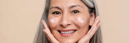 Eine lächelnde Frau mit grauen Haaren hat kleine Mengen Creme auf beiden Wangen aufgetragen. Ihre Hände sind sanft unter ihrem Gesicht positioniert, während sie in die Kamera schaut. Der Hintergrund ist in einem neutralen Beige gehalten.